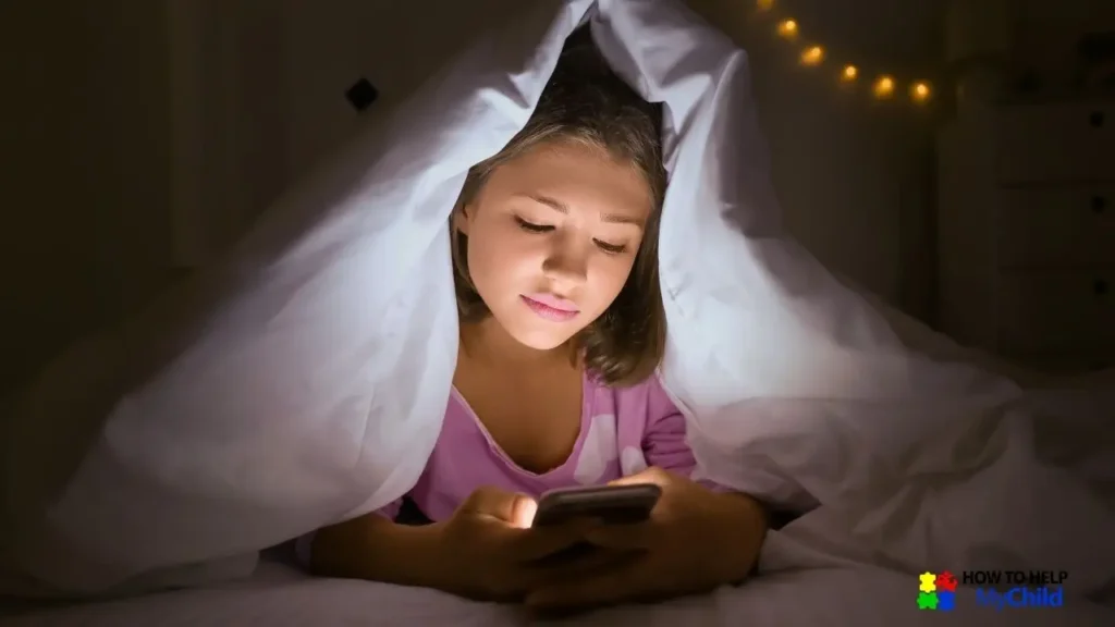 A young girl in pink pajamas lies under a blanket in a dimly lit bedroom, engrossed in her phone screen. The soft glow of fairy lights in the background contrasts with the bright screen light, illustrating the challenge of limiting screen time before bed. Reducing screens before sleep is an important part of a structured bedtime routine for children with autism and ADHD. The "How to Help My Child" logo is visible in the corner.